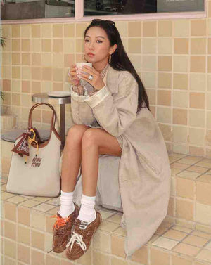 High-angle on-feet shot of brown suede New Balance shoes on tiled stairs, paired with slouched white crew socks and featuring double laces in orange and off-white alongside Miu Miu tongue branding.