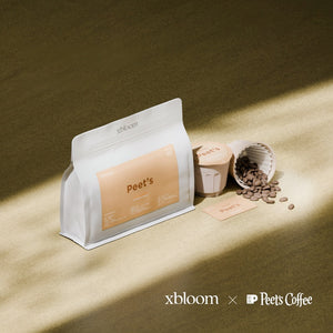 A white bag of Peet's Coffee next to individual XBloom xpods coffee capsules and loose roasted beans on a textured surface, showcasing the 'xbloom x Peet's Coffee' collaboration logo.
