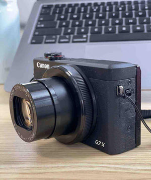 Front view of the compact black Canon PowerShot G7 X Mark III sitting on a desk in front of a laptop keyboard.