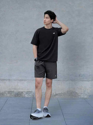Full-length lifestyle shot of a man standing against a grey textured wall, wearing a black New Balance t-shirt, dark shorts, white socks, and grey New Balance Fresh Foam X running shoes.
