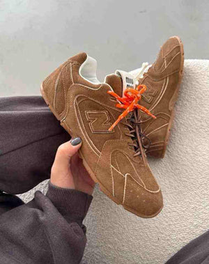 New Balance shoes x Miu Miu collaboration held in hand, displaying brown suede upper construction, exposed stitching, bright orange laces, and co-branded tongue logo.