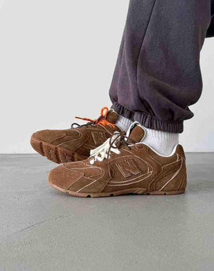 On-feet side profile of brown suede New Balance shoes styled with dark grey sweatpants, featuring off-white and brown double laces, contrast stitching, and white socks.