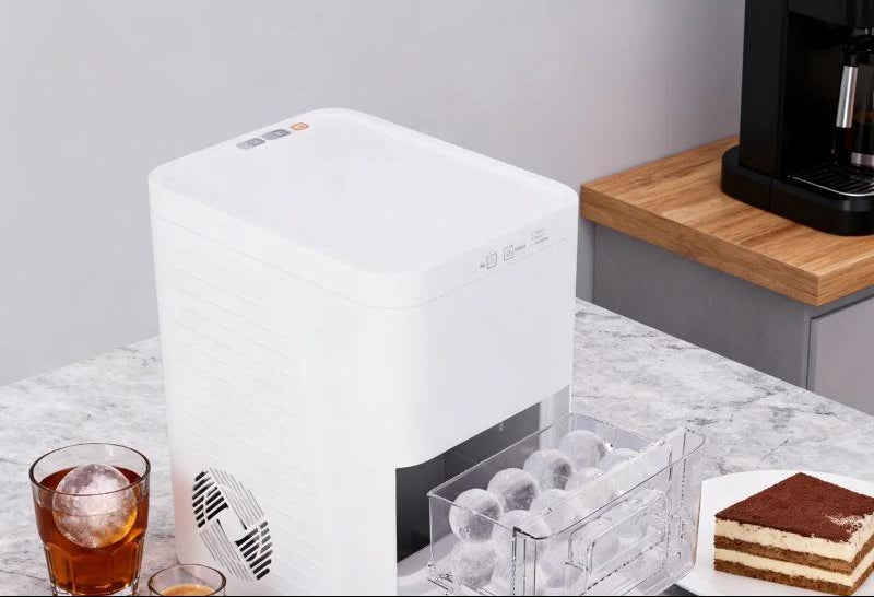 A compact white ice maker machine operating on a kitchen counter next to iced drinks and cakes.